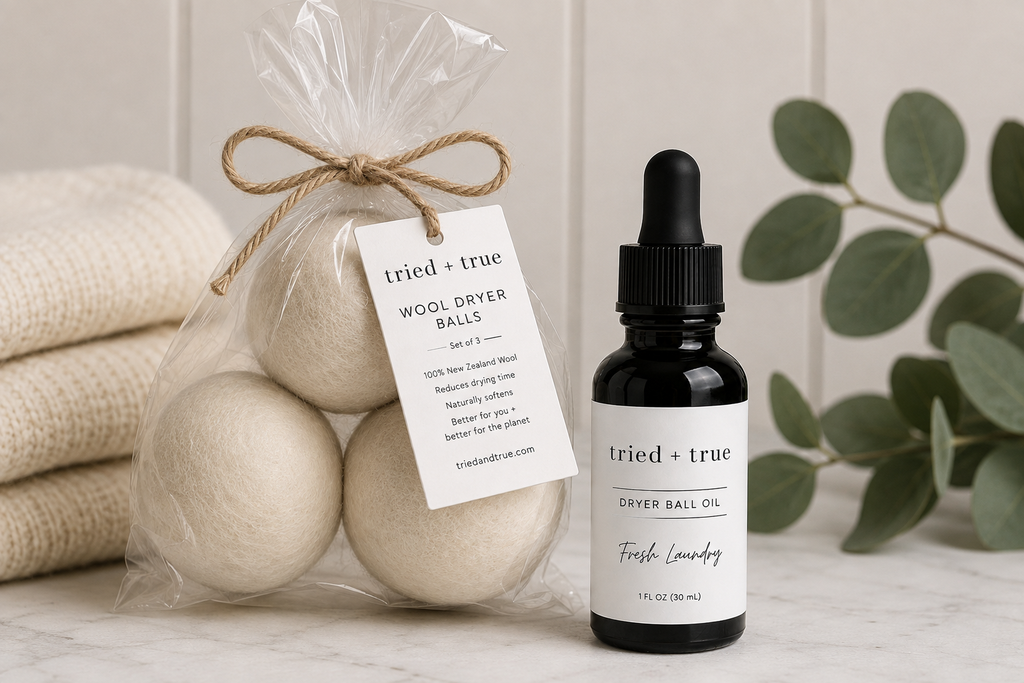 A small black dropper bottle labeled tried + true Dryer Ball Oil Fresh Laundry stands next to a clear bag of three wool dryer balls tied with twine and a hanging product tag. The scene is styled on a light marble surface with folded beige fabric and green eucalyptus leaves in the background.