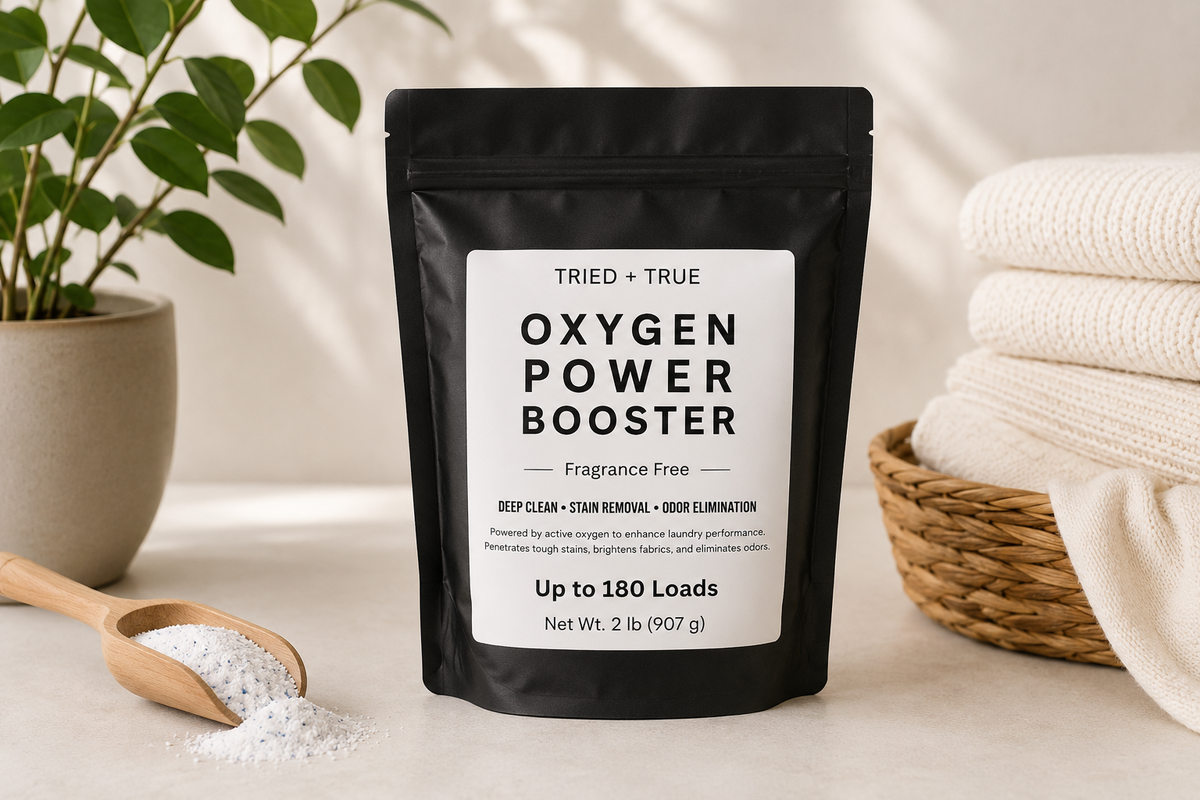 A black stand-up pouch labeled 'TRIED + TRUE OXYGEN POWER BOOSTER' stands on a light surface, with white powder spilled in a wooden spoon nearby. A potted plant is on the left and folded white towels in a basket are on the right, creating a clean, home-like setting.