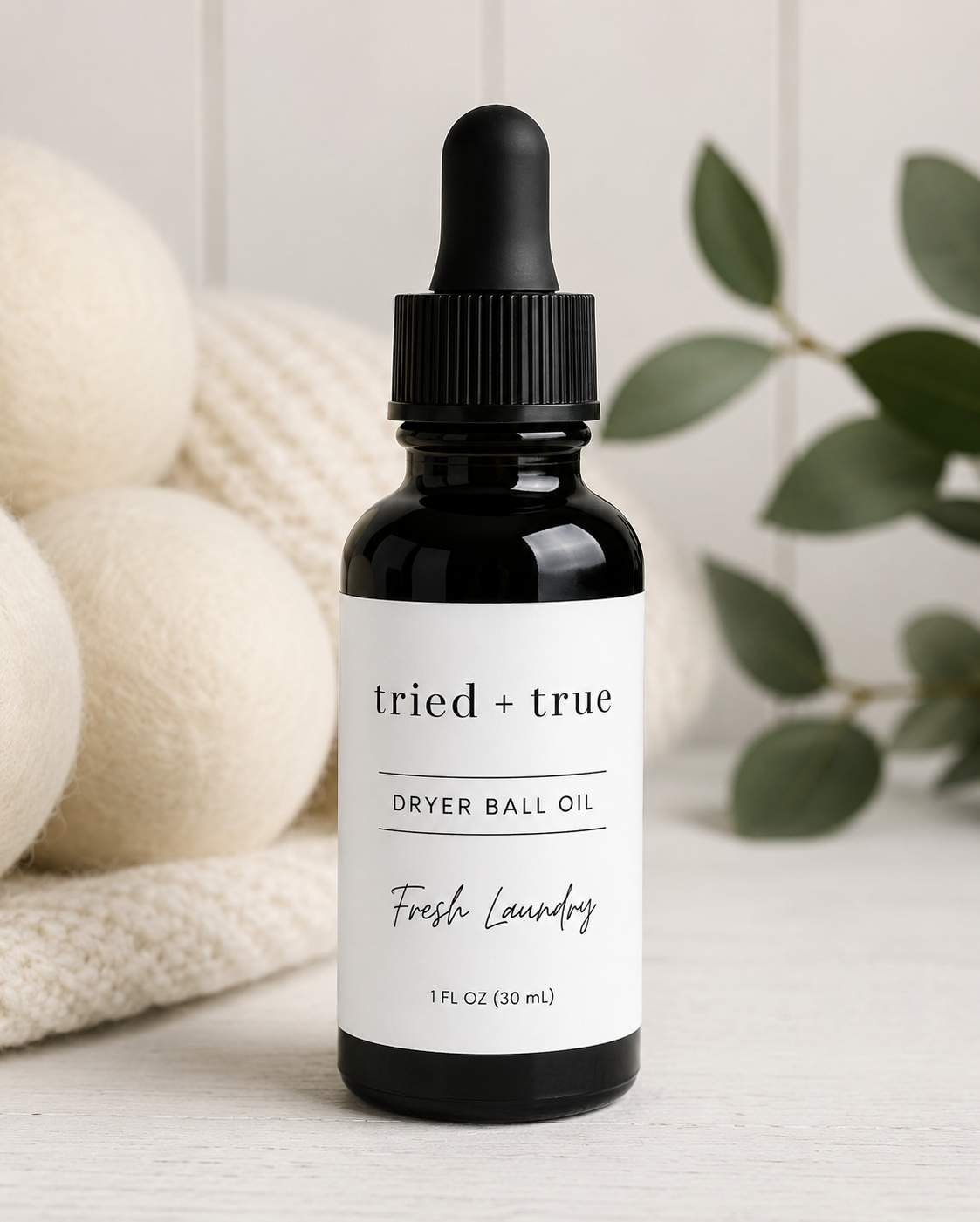 A 1 fl oz black glass dropper bottle of tried + true Dryer Ball Oil in the Fresh Laundry scent, styled on a white surface with a cream knit blanket and green leaves in the background.